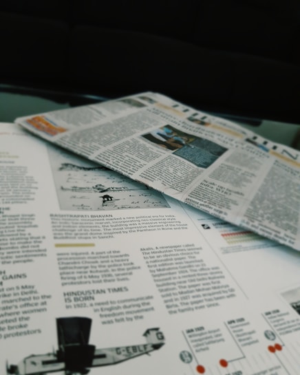 Newspapers and magazines are spread out, with text and images visible on the pages. One page has an article about historical topics, including a photo of an aircraft and a section discussing the inauguration of a political monument.