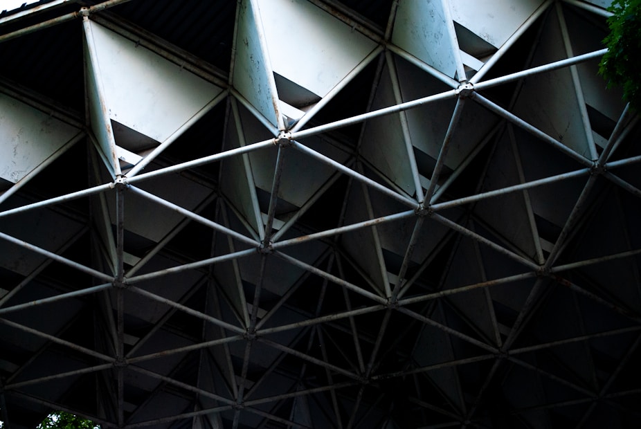 An intricate structure composed of metal bars forming geometric patterns. The lattice-like framework creates a repetitive triangular and diamond-shaped design. The lighting is dim with shadows emphasizing the depth and complexity of the construction.