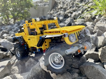 A brightly colored yellow LEGO model of a Rubicon off-road vehicle is positioned on a set of large, uneven gray rocks. The vehicle features detailed elements like large tires, headlights, and a front bumper, with the brand name clearly marked on the side. Sunlight illuminates the scene, highlighting both the vehicle and the rocky terrain. Sparse green vegetation can be seen in the background, adding a touch of nature to the rugged setting.