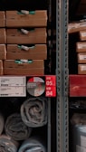 Several cardboard boxes labeled 'LYCKSELE' are stacked on shelves alongside rolled up plastic-wrapped items. The shelving rack features labels and tags written in multiple languages, including English and possibly Chinese. The area appears to be a warehouse or store aisle.