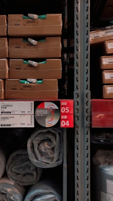 Several cardboard boxes labeled 'LYCKSELE' are stacked on shelves alongside rolled up plastic-wrapped items. The shelving rack features labels and tags written in multiple languages, including English and possibly Chinese. The area appears to be a warehouse or store aisle.