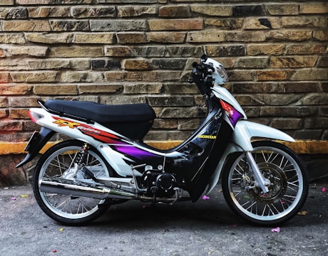 A motorcycle is parked against a textured stone wall. The bike has a black seat, with a sleek body painted in white, black, and purple, and features a colorful decal. The wall has various shades of brown and beige, and the ground is concrete with small scattered pink petals.