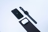 A neatly arranged set of electronic devices on a clean white background, including a black smartphone, a black smartwatch, and a pair of white wireless earbuds with their case placed on a black mat.