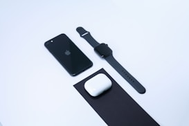A neatly arranged set of electronic devices on a clean white background, including a black smartphone, a black smartwatch, and a pair of white wireless earbuds with their case placed on a black mat.
