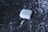 A white wireless earbud charging case is floating against a metallic, reflective background. Attached to the case is a charging cable with a connector visible in the foreground. The green light on the case indicates it is powered on.