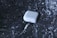 A white wireless earbud charging case is floating against a metallic, reflective background. Attached to the case is a charging cable with a connector visible in the foreground. The green light on the case indicates it is powered on.