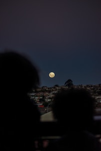 A large moon is visible in the night sky above a silhouette of buildings and trees on the horizon. The scene captures a quiet, residential area with soft lighting. Two figures are faintly visible in the foreground, adding depth and a sense of scale.