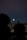A large moon is visible in the night sky above a silhouette of buildings and trees on the horizon. The scene captures a quiet, residential area with soft lighting. Two figures are faintly visible in the foreground, adding depth and a sense of scale.