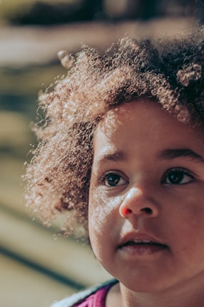 A close-up portrait of a child with a natural, curious expression in a sunlit park.