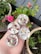 A hand holds four small round candles embedded with dried flower petals and herbs. In the background, there are potted plants with pink roses and green foliage, set against a patio or garden scene with a wooden deck.
