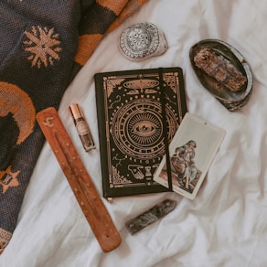 An arrangement of mystical and spiritual items on a soft white fabric. A notebook with intricate designs featuring an eye symbol, alongside a tarot card depicting a seated figure. A wooden incense holder, a crystal, and a small bottle of essential oil or perfume are nearby. A decorative stone with black patterns and a bundle of dried herbs rest on a small black dish. A woven fabric with sun and moon designs is draped in the background.