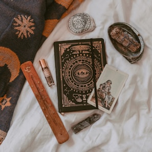 An arrangement of mystical and spiritual items on a soft white fabric. A notebook with intricate designs featuring an eye symbol, alongside a tarot card depicting a seated figure. A wooden incense holder, a crystal, and a small bottle of essential oil or perfume are nearby. A decorative stone with black patterns and a bundle of dried herbs rest on a small black dish. A woven fabric with sun and moon designs is draped in the background.