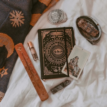 An arrangement of mystical and spiritual items on a soft white fabric. A notebook with intricate designs featuring an eye symbol, alongside a tarot card depicting a seated figure. A wooden incense holder, a crystal, and a small bottle of essential oil or perfume are nearby. A decorative stone with black patterns and a bundle of dried herbs rest on a small black dish. A woven fabric with sun and moon designs is draped in the background.