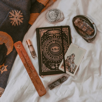 An arrangement of mystical and spiritual items on a soft white fabric. A notebook with intricate designs featuring an eye symbol, alongside a tarot card depicting a seated figure. A wooden incense holder, a crystal, and a small bottle of essential oil or perfume are nearby. A decorative stone with black patterns and a bundle of dried herbs rest on a small black dish. A woven fabric with sun and moon designs is draped in the background.