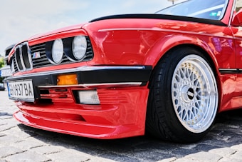 A close-up view of a red sports car with a distinctive front bumper and BBS alloy wheels. The car features circular headlights and a license plate with alphanumeric characters. The image focuses on the front side, showcasing the car's sleek design and vibrant paint job.