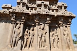 Intricate stone carvings depict a series of human figures in traditional attire, framed by ornate columns. The structure showcases detailed craftsmanship with multiple layers of sculpted figures, highlighting a historical or cultural narrative.
