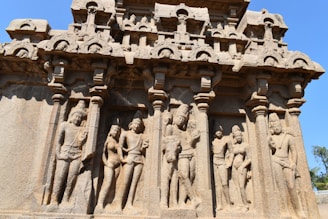 Intricate stone carvings depict a series of human figures in traditional attire, framed by ornate columns. The structure showcases detailed craftsmanship with multiple layers of sculpted figures, highlighting a historical or cultural narrative.