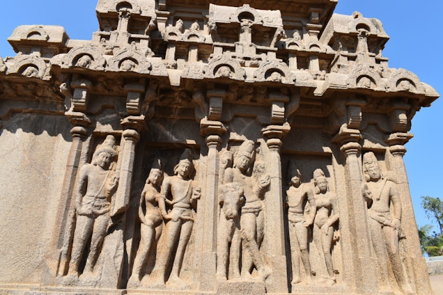 Intricate stone carvings depict a series of human figures in traditional attire, framed by ornate columns. The structure showcases detailed craftsmanship with multiple layers of sculpted figures, highlighting a historical or cultural narrative.