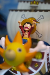 A cartoon figure with an excited expression, wearing a straw hat and a red and yellow outfit, holds its arms up in celebration. The figure is in front of a ship with a large sail, adorned with a skull and crossbones design. There's a bright yellow, sun-shaped figure in the foreground, slightly blurred.