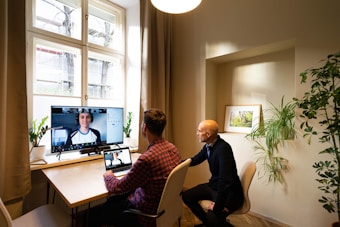 Two individuals are in a well-lit room with a window on the left side. One person, wearing a checkered shirt, is seated at a desk with a laptop displaying a video call. A larger screen on the desk also shows the same video call. The other person, dressed in a dark sweater, is sitting beside the first individual, facing the screen. The room has a bright, natural feel, with several plants placed around the space and a framed picture on the wall.