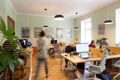A busy office environment with multiple people working at shared desks. Laptops and computers are actively being used, and there are plants and personal items on the desks. The room has light green walls with artistic posters, wooden flooring, and hanging lights. Natural light enters through the large windows, creating a bright atmosphere. A person is walking, creating a dynamic atmosphere.