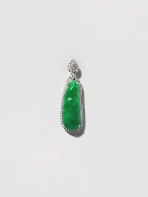 A pendant featuring an elongated green gemstone set within a detailed silver or platinum setting, encrusted with small, sparkling stones around the perimeter. The pendant is suspended from an intricate floral-shaped bail, also adorned with sparkling stones.
