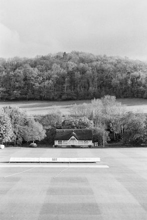 A black and white photograph of a rural scene with a small house quaintly nestled among trees at the edge of a woodland area. The house has a thatched roof and is surrounded by bushes and trees. In the foreground, there is a large, open, flat area that appears to be a sports field or lawn, with a covered structure near the house.