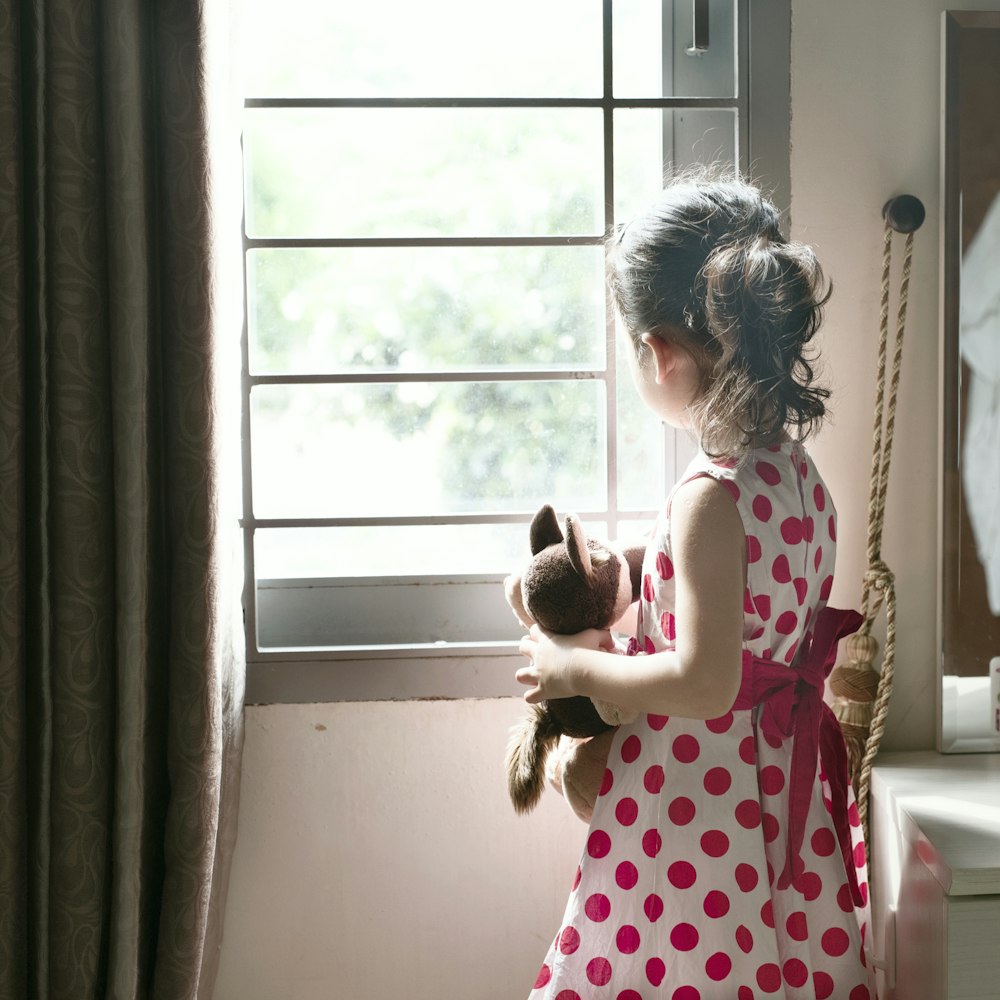 빨간색과 흰색 물방울 무늬 드레스를 입은 소녀가 창문 옆에 서 있다