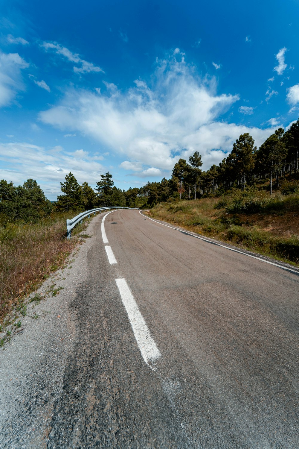 Route en béton gris entre un champ d’herbe verte sous un ciel bleu et des nuages blancs pendant la journée