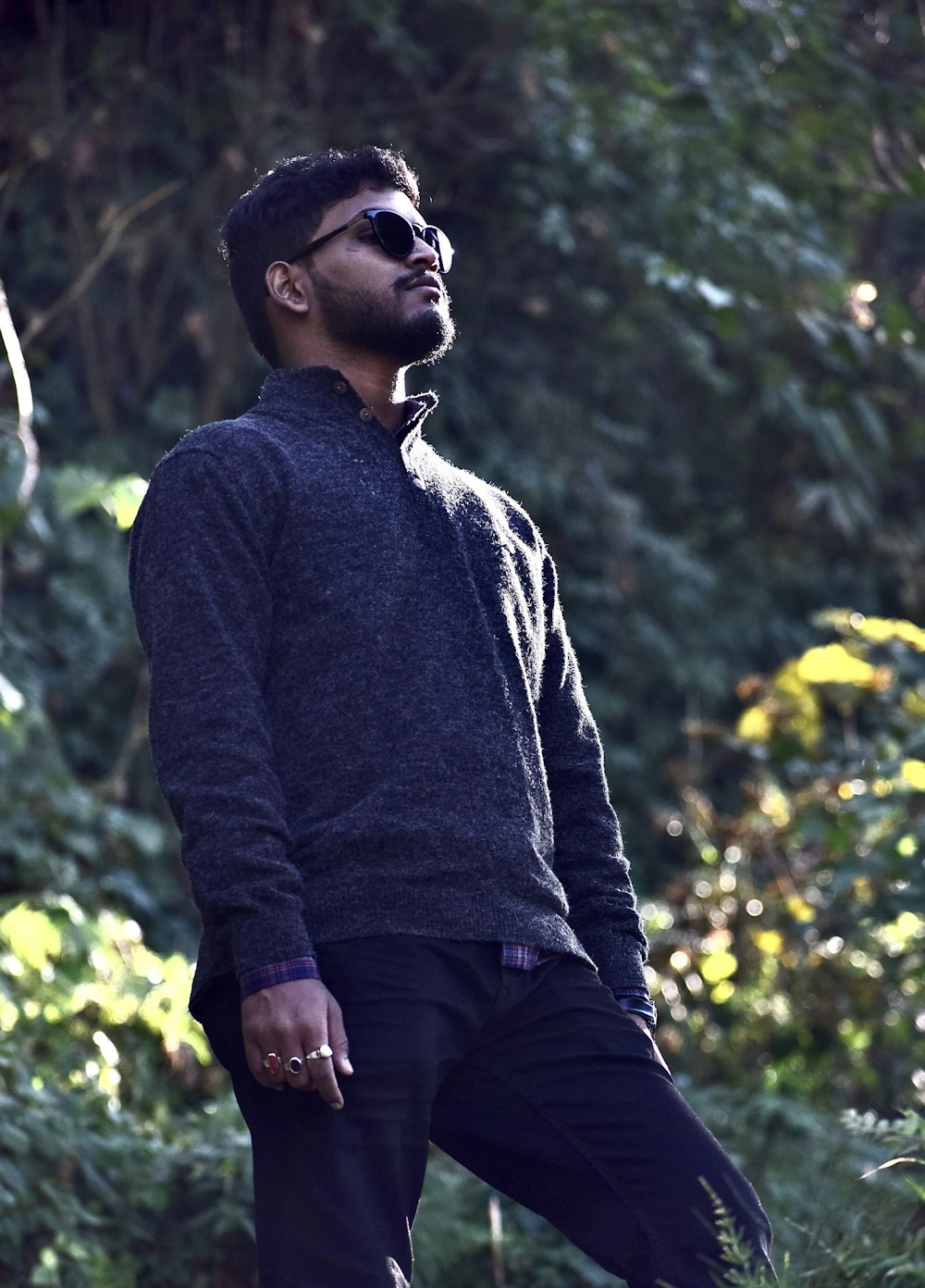 man in gray sweater and black pants wearing black sunglasses standing near green trees during daytime