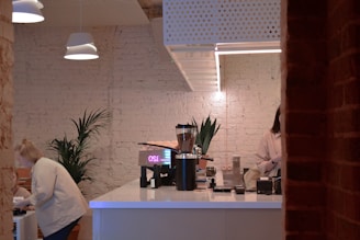 A modern café with a white brick wall and wooden accents. The counter area includes a coffee machine, grinder, and other coffee-related equipment. Potted plants are placed for decoration. Two people, one customer and one barista, are engaging in their activities within the café.
