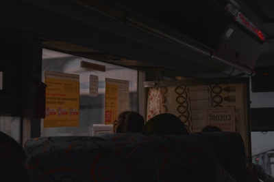 The interior of a dimly lit bus with patterned seats and signage on the windows. Passengers are sitting, with some partially visible. The windows display several information cards in a language similar to Russian and an emergency exit sign.