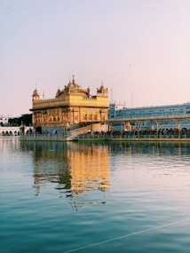 A beautifully ornate golden temple is prominently reflected in a calm body of water. The architecture features intricate carvings and multiple domes, set against a serene sky and surrounded by a crowd of visitors.