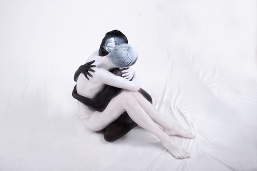 Two people are intertwined in a sitting embrace, with contrasting body paint. One person is painted entirely in black, and the other in white, against a neutral backdrop, highlighting the contrast and unity.