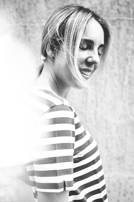A black and white photograph of a person with light hair, wearing a striped shirt. The image captures the individual smiling with eyes closed, creating a serene and content mood. The background is slightly blurred, drawing focus to the subject.