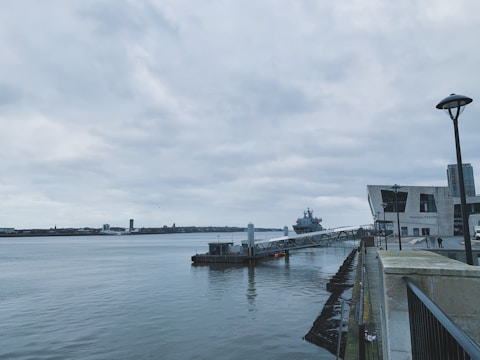 A waterfront scene featuring a large body of water with a city skyline in the distance. A substantial modern ferry terminal is visible on the right, and a docked naval ship can be seen nearby. The sky is overcast, and the setting has a calm, serene atmosphere.