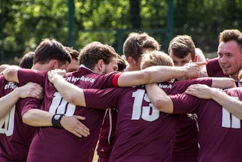 A group of soccer players wearing maroon jerseys are huddled together with arms around each other, displaying camaraderie and team spirit. The background shows a green, outdoor setting.