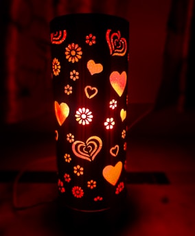 A cylindrical lamp emits a warm red glow, adorned with heart and flower cutouts that allow light to shine through. The pattern of hearts and flowers creates a cozy, romantic atmosphere.