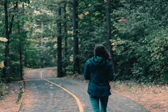 woman in green jacket walking on pathway between trees during daytime