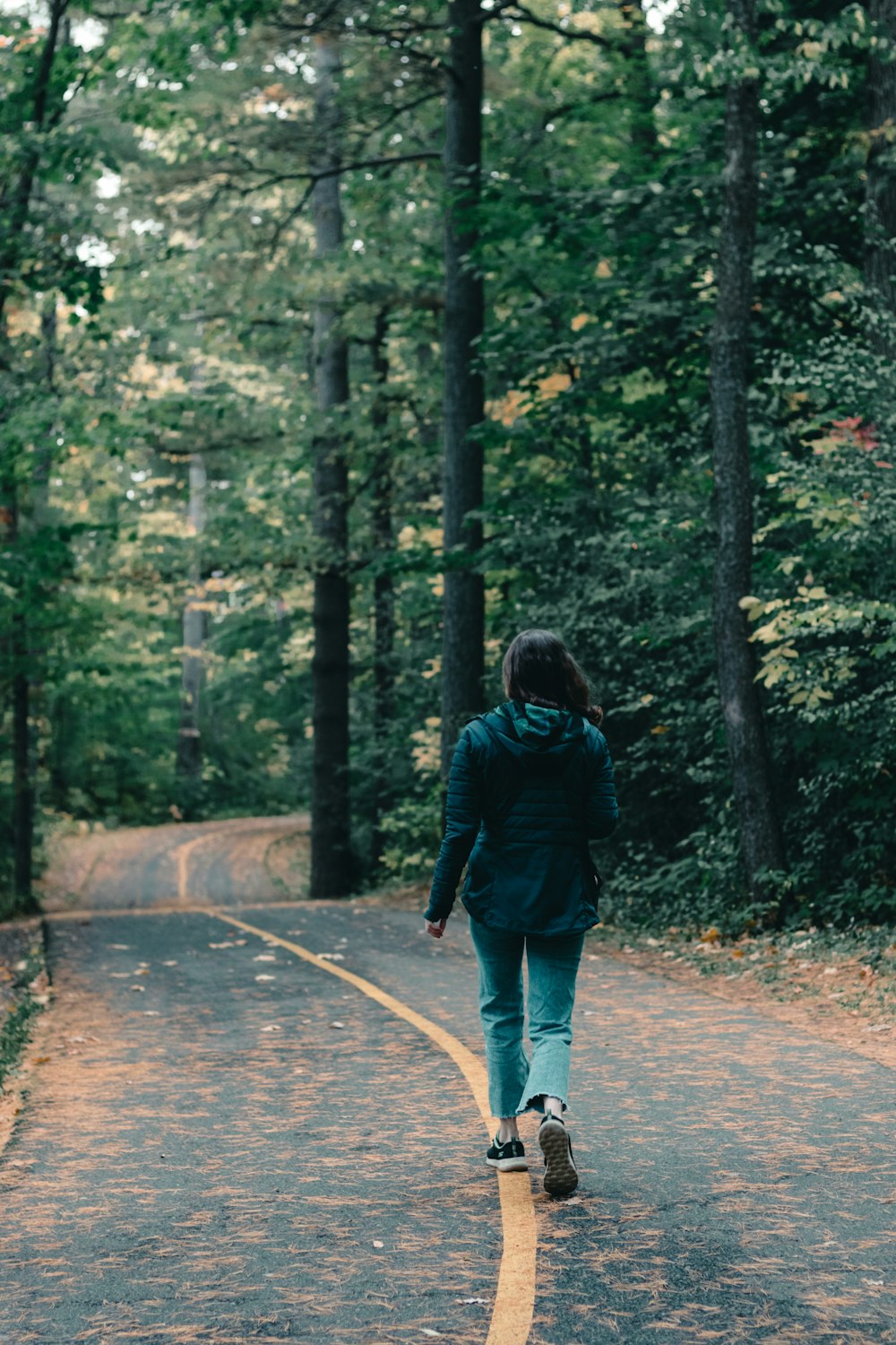 Mujer con chaqueta negra caminando por el camino entre los árboles durante el día