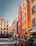 A vibrant city square with colorful buildings featuring red, orange, and yellow facades. People are dining at outdoor tables of a trattoria along the street. The cobblestone pavement and historic architecture suggest a European setting.
