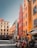 A vibrant city square with colorful buildings featuring red, orange, and yellow facades. People are dining at outdoor tables of a trattoria along the street. The cobblestone pavement and historic architecture suggest a European setting.