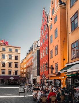 A vibrant city square with colorful buildings featuring red, orange, and yellow facades. People are dining at outdoor tables of a trattoria along the street. The cobblestone pavement and historic architecture suggest a European setting.