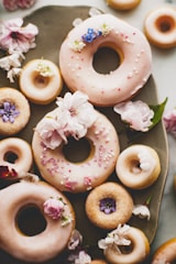A collection of donuts adorned with delicate pink icing and colorful sprinkles is set on a rustic platter. Various edible flowers, including small blue and purple blooms, add a whimsical touch. The atmosphere is enhanced by cherry blossom petals scattered across the surface, complementing the pastel color scheme.