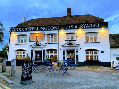 A traditional pub with a white exterior, displaying the name 'Duke of Wellington' and the year 'c.1516 Ryarsh' in black signage. Warm yellow lights illuminate the sign and front of the building. The pub has multiple windows with white frames and a pitched roof with dark shingles. In front, there are several blue metal outdoor tables and chairs set on a paved area, along with a blackboard sign advertising Christmas party bookings. A few potted plants are placed near the entrance.