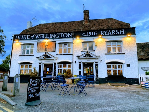 A traditional pub with a white exterior, displaying the name 'Duke of Wellington' and the year 'c.1516 Ryarsh' in black signage. Warm yellow lights illuminate the sign and front of the building. The pub has multiple windows with white frames and a pitched roof with dark shingles. In front, there are several blue metal outdoor tables and chairs set on a paved area, along with a blackboard sign advertising Christmas party bookings. A few potted plants are placed near the entrance.