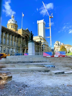 A historic building with a dome and flag stands behind a statue on a pedestal in a public square. A large crane is visible over a partially constructed building, and construction materials are scattered in the foreground.