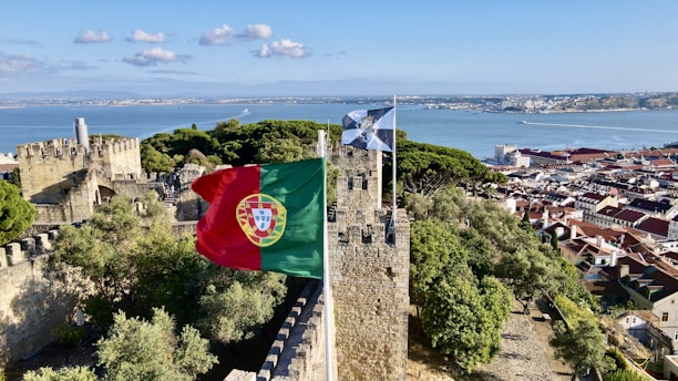 A historic stone castle overlooking a scenic bay with lush green trees. The Portuguese flag flutters in the wind atop the castle walls. In the background, there is a panoramic view of a coastal city with red roofed buildings and a serene blue ocean.