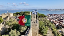 A historic stone castle overlooking a scenic bay with lush green trees. The Portuguese flag flutters in the wind atop the castle walls. In the background, there is a panoramic view of a coastal city with red roofed buildings and a serene blue ocean.