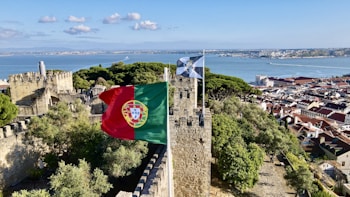 A historic stone castle overlooking a scenic bay with lush green trees. The Portuguese flag flutters in the wind atop the castle walls. In the background, there is a panoramic view of a coastal city with red roofed buildings and a serene blue ocean.